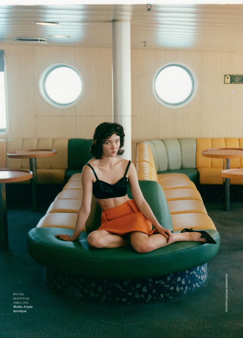 A young brunette woman in a black bra top and orange skirt sits on a green and blue cushioned platform in an indoor setting with round windows.