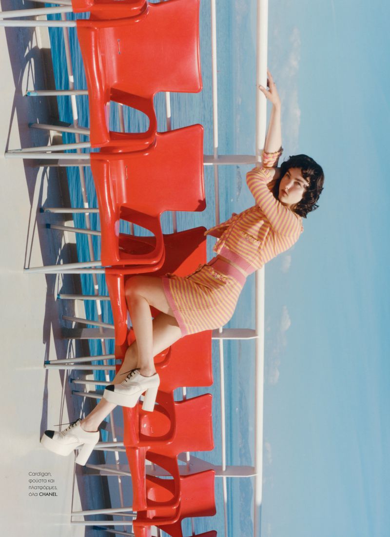 Brunette woman in stylish, orange, knitted dress and platform shoes poses on a row of red chairs on the deck with ocean backdrop.