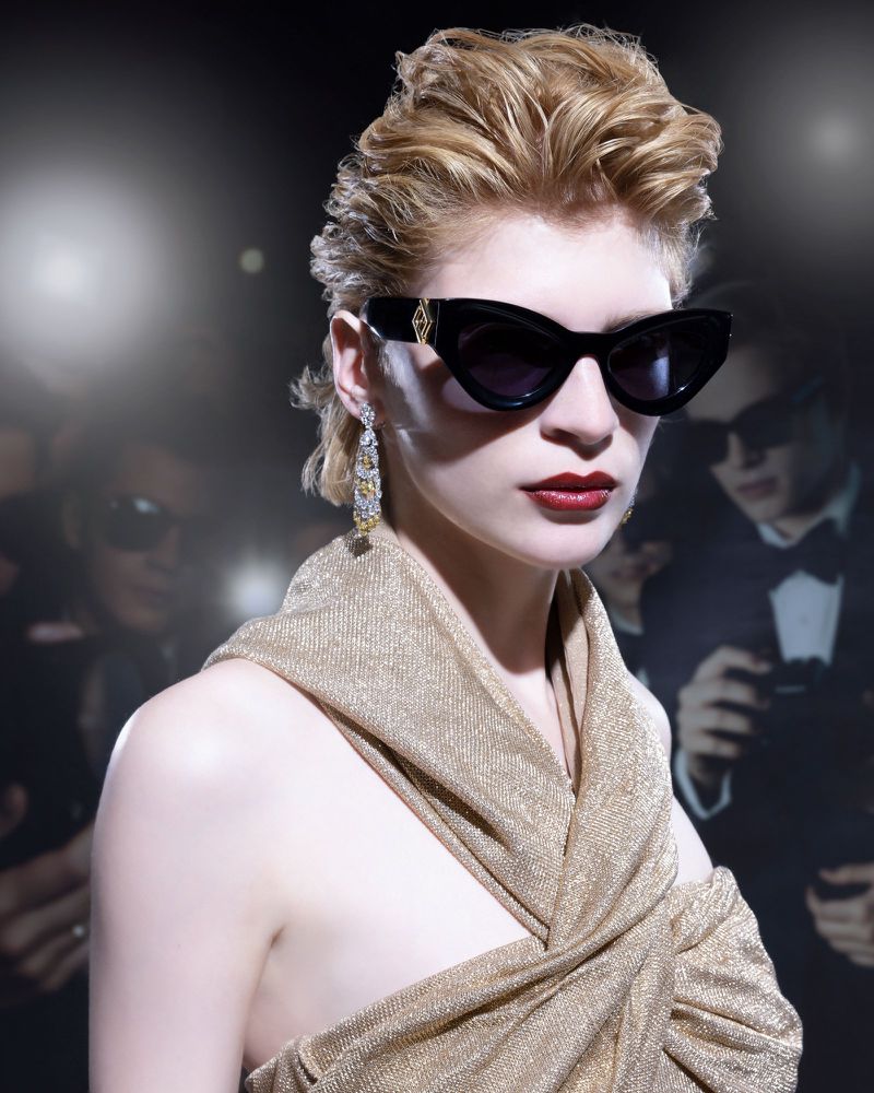 Woman in sunglasses and draped gold fabric poses in front of paparazzi flashbulbs, evoking celebrity and entertainment.