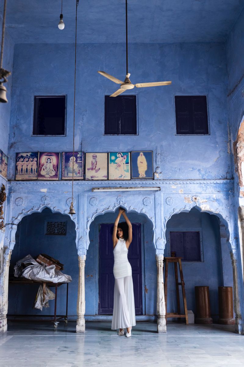 A woman in a white outfit poses in a blue-toned indoor space with arches, vintage posters, and a rustic aesthetic.
