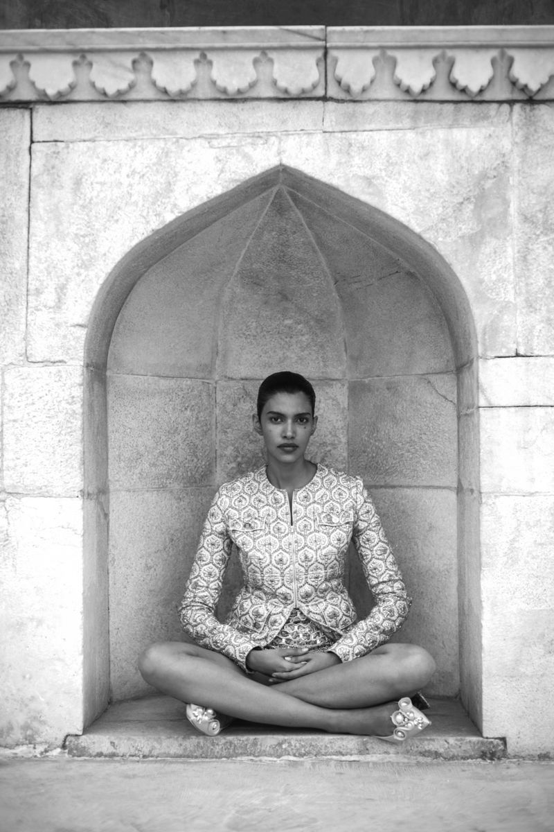 Monochrome shot of an adult female in patterned jacket sitting in lotus position within a stone archway, fingers interlaced, serene expression.