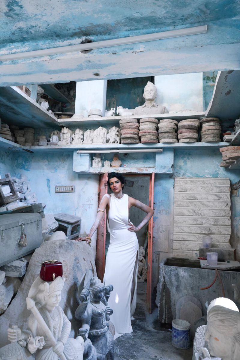 Woman in elegant white dress poses inside a sculpture workshop, surrounded by stone artwork and blue walls, under natural light.
