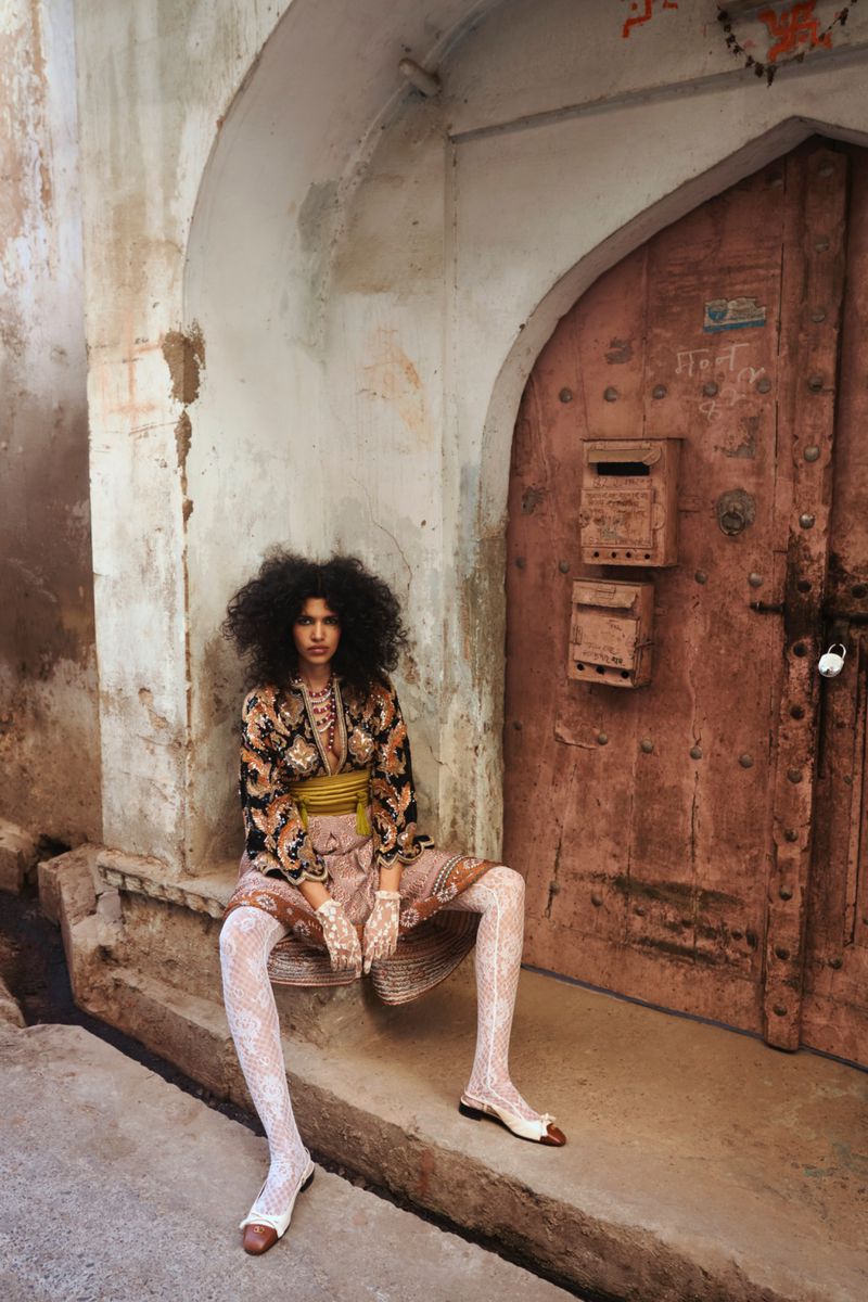 An adult female with dark, curly hair poses while sitting in front of an arched doorway with a weathered, pink door and mailboxes.