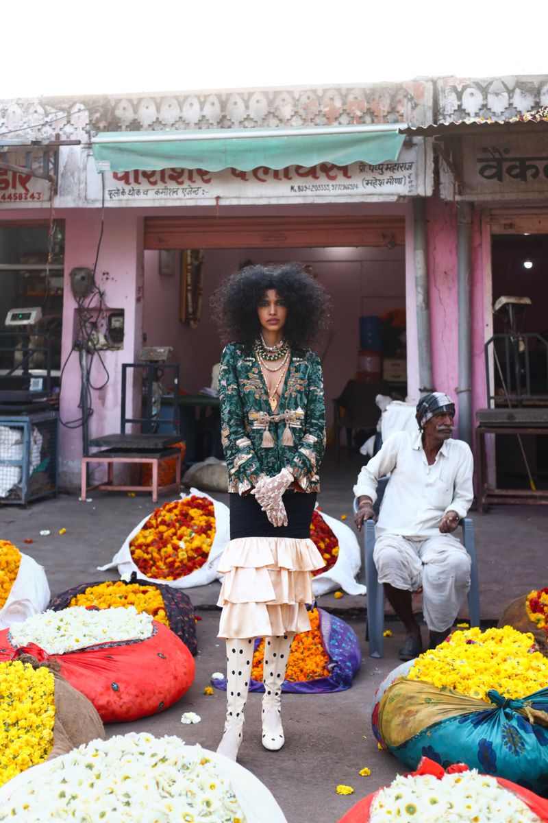 Fashion model poses outdoors near flower stalls, wearing a green and gold jacket, black skirt, and polka dot tights, with an afro hairstyle.