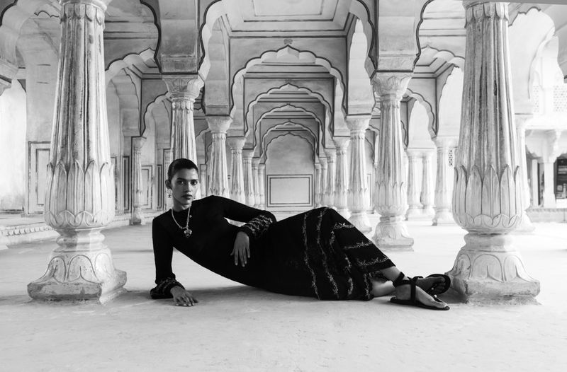 Monochrome fashion shot: a model in a black dress poses between ornate pillars in an arched hallway, exuding elegance and style.