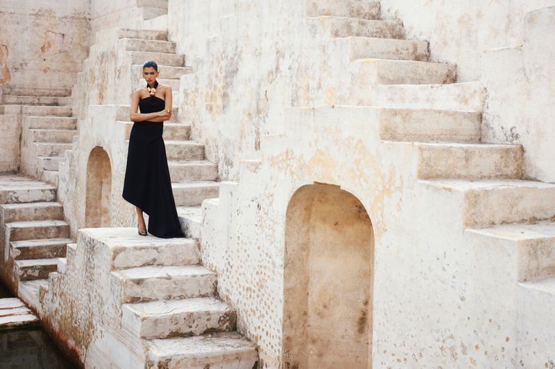 Fashion shot of a solitary female model wearing a black gown, posing on outdoor stone steps with an ancient design and neutral palette.