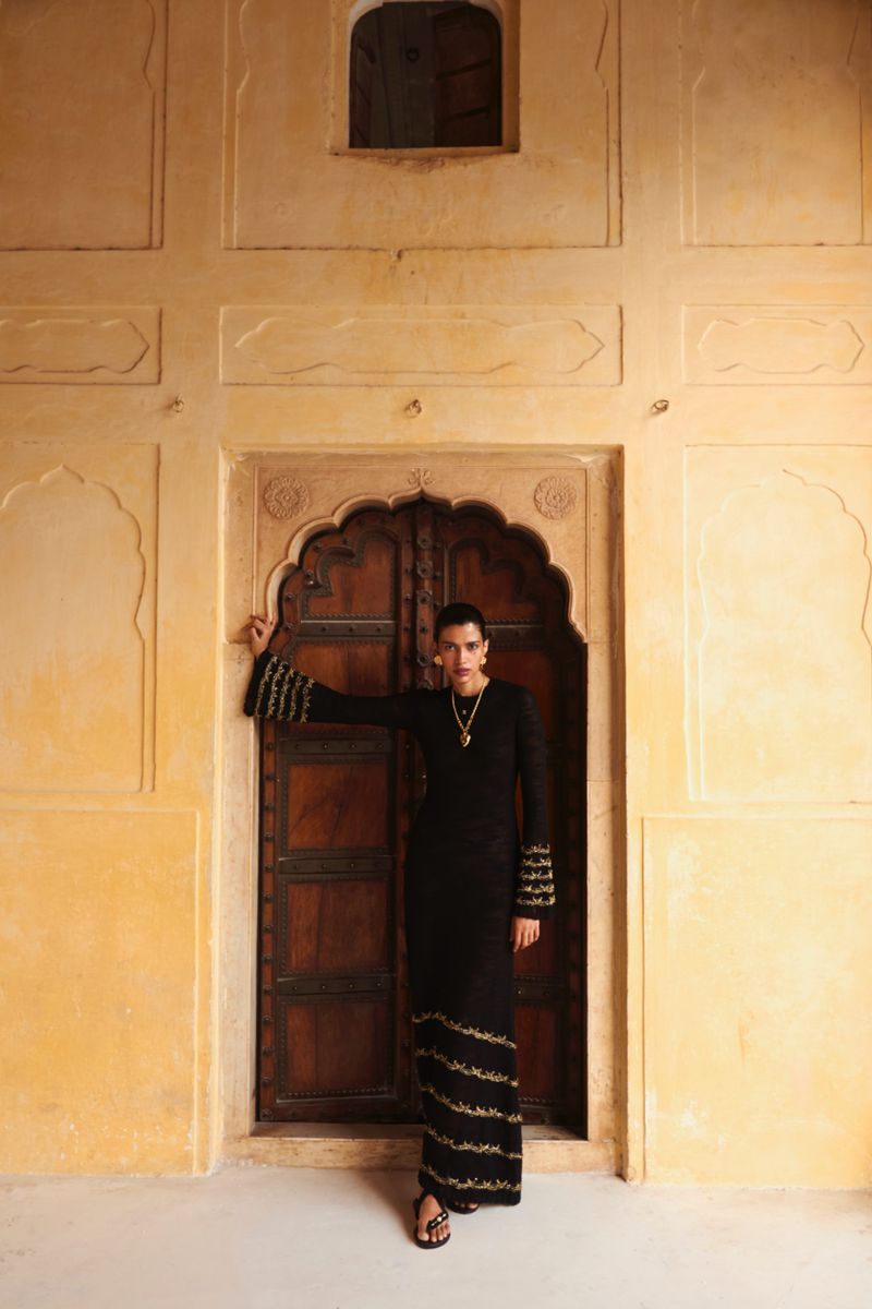 A woman in a black dress with gold embroidery poses in front of an ornate wooden door and a textured, pale orange wall.