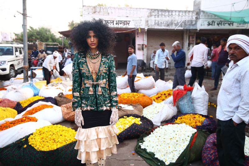 Fashion shot featuring a woman with an afro hairstyle in an ornate jacket surrounded by bags of colorful flowers in an outdoor market.