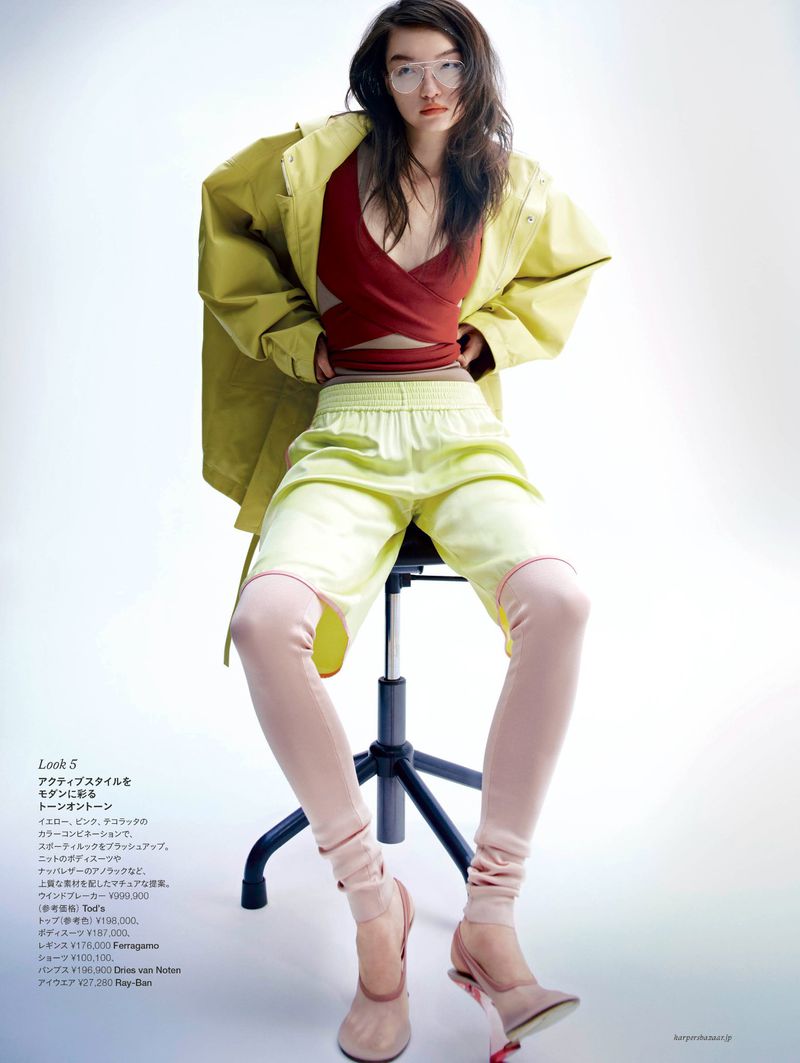 A slender young woman wearing glasses is sitting on a black chair, wearing a red crop top, a yellow jacket, and pale pink leggings.