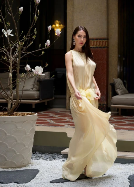 Woman in flowing yellow dress poses with flowers in an indoor courtyard with tile floor and neutral seating. Fashion and beauty photography.