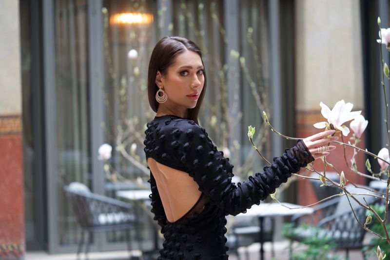 Brunette woman in black dress poses in courtyard with blossoming branches. Fashion editorial shot in soft, diffused light.