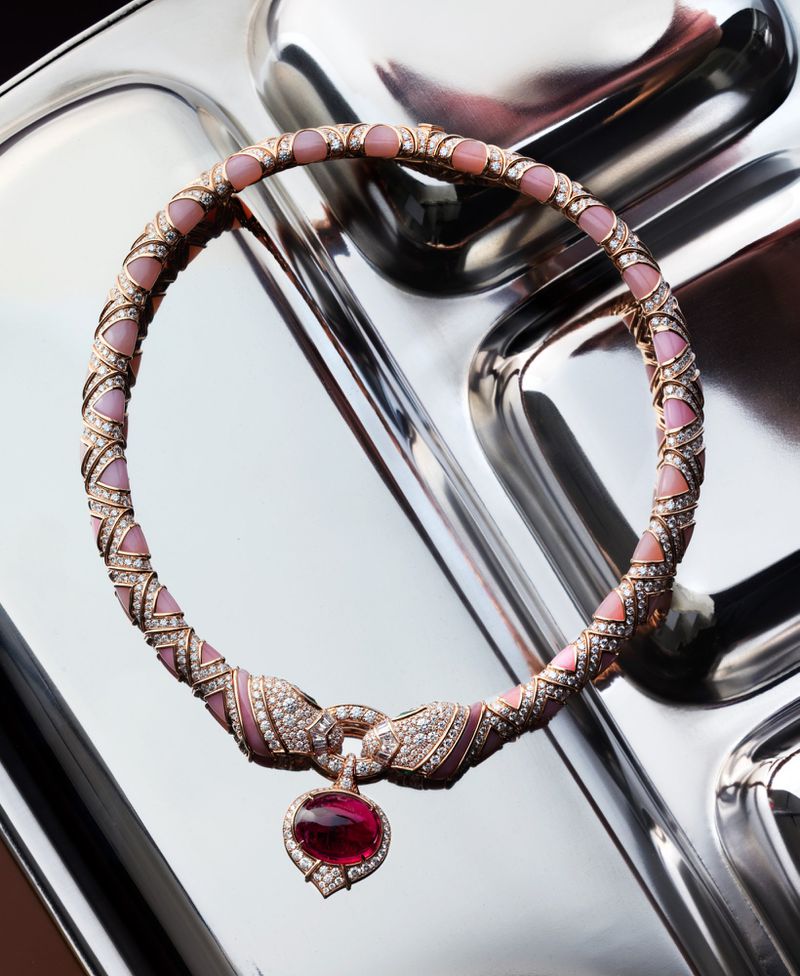 High-angle, close-up of a luxurious pink and gold gemstone necklace with a ruby pendant displayed on a shiny, reflective silver tray.