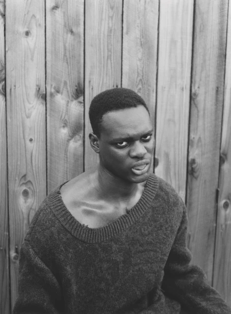 Monochrome portrait of a Black man in a sweater against a wood panel backdrop, showcasing a contemplative yet intense gaze and serious expression.