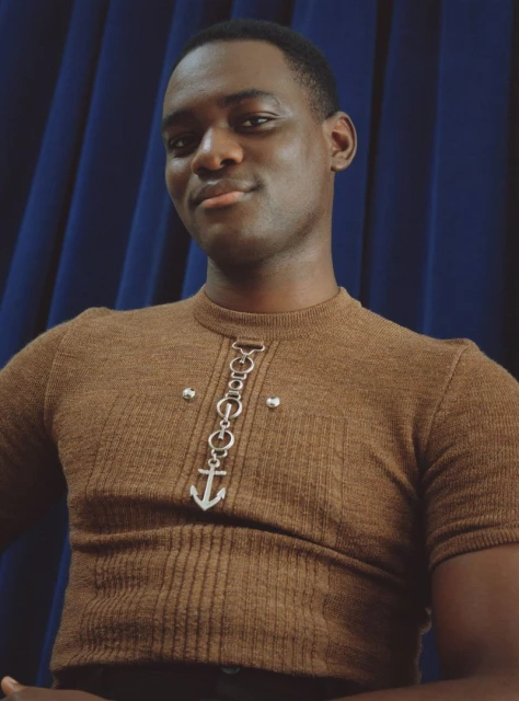 Portrait of a Black man wearing a brown ribbed shirt with a silver anchor pendant against a blue curtain background, indoor studio setting.