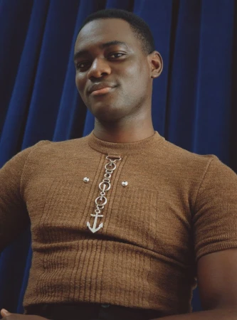 Portrait of a Black man wearing a brown ribbed shirt with a silver anchor pendant against a blue curtain background, indoor studio setting.