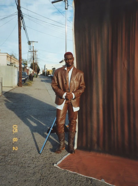 Full body shot of a Black man in a brown leather suit posing in front of a brown backdrop on an outdoor set on a sunny day.