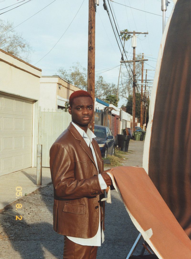 An African American man in a brown leather suit poses outdoors on a city street next to photography equipment under a cloudy sky.