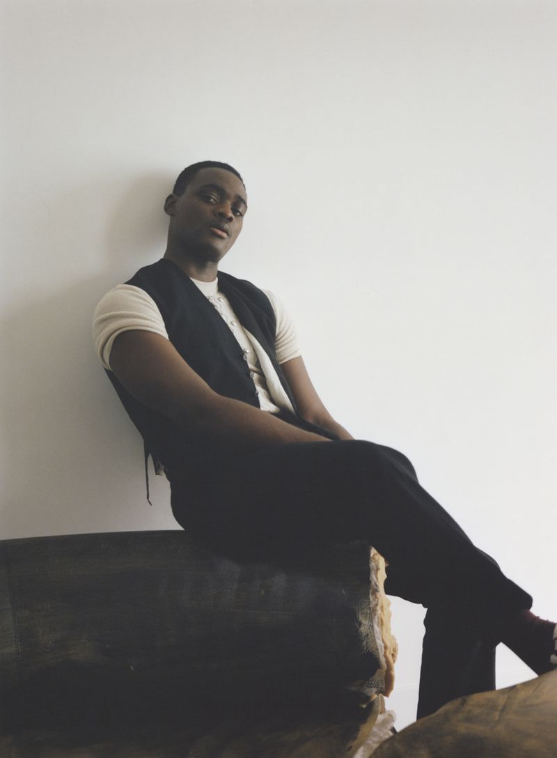 An African American man poses indoors, wearing a vest and light shirt while sitting on a wooden surface against a white wall backdrop.