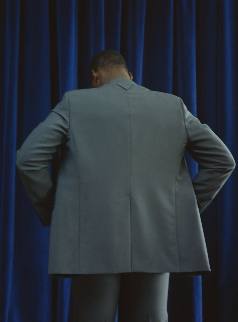 Back view of an adult male in a gray suit standing in front of a blue curtain in a studio setting, facing away from the camera.