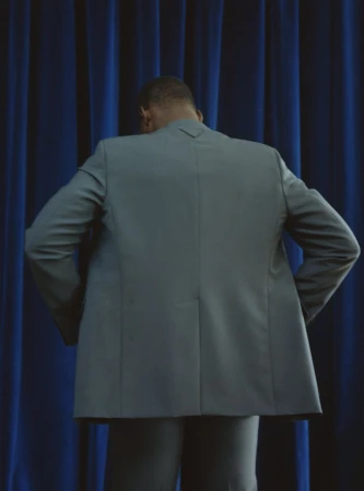 Back view of an adult male in a gray suit standing in front of a blue curtain in a studio setting, facing away from the camera.