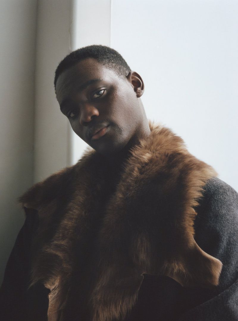 Close-up of an adult Black male wearing a fur stole and gray coat. He is indoors with diffused natural lighting.