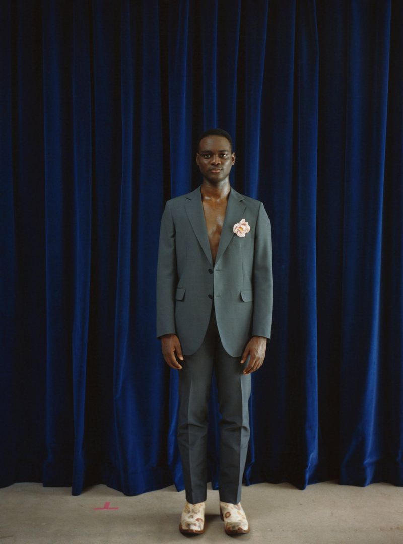 A Black man in a teal suit stands centered against a draped dark blue curtain backdrop, wearing floral shoes, and gazing forward.