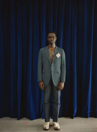 A Black man in a teal suit stands centered against a draped dark blue curtain backdrop, wearing floral shoes, and gazing forward.