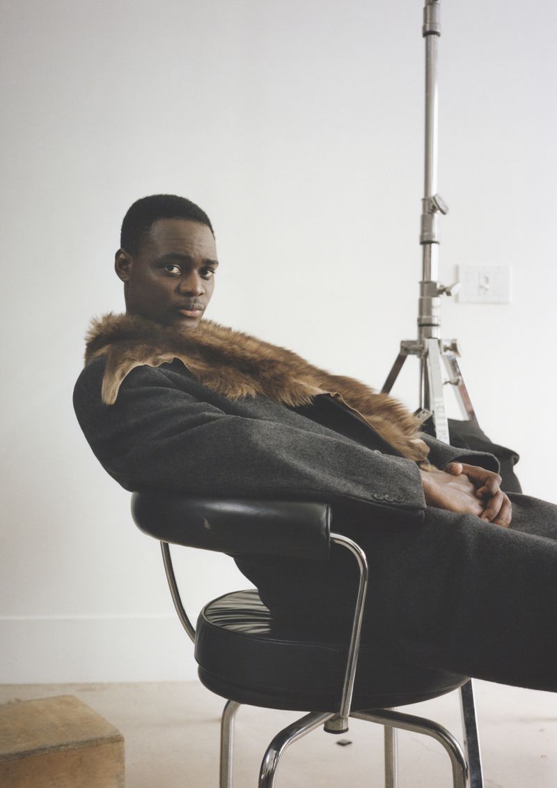 Portrait of an adult male in a gray coat with a fur collar, sitting in a black chair in a photo studio with lighting equipment.