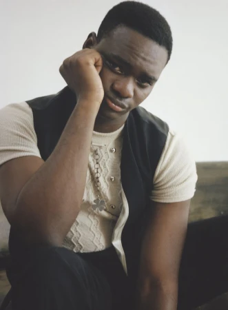 Portrait of a dark-skinned man with short hair in a vest and short-sleeved shirt, hand near his face, gazing into the distance.