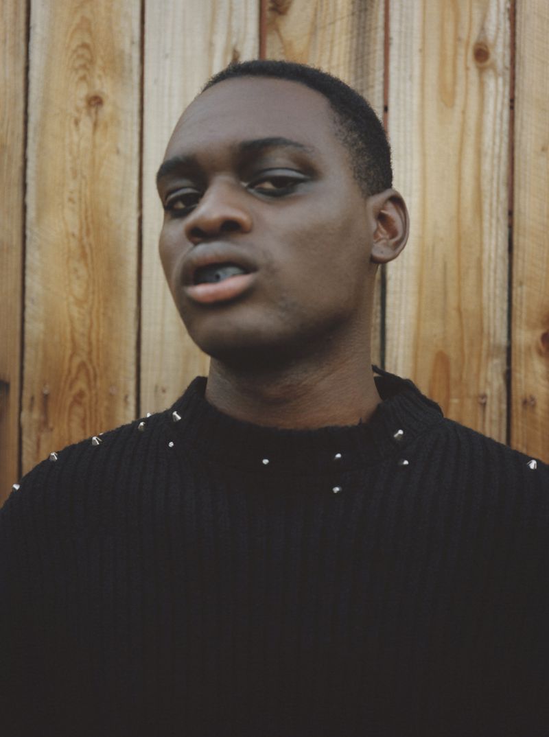 Close-up of a Black male wearing a black studded sweater posing outdoors against a wood background, shot in portrait format with natural lighting.