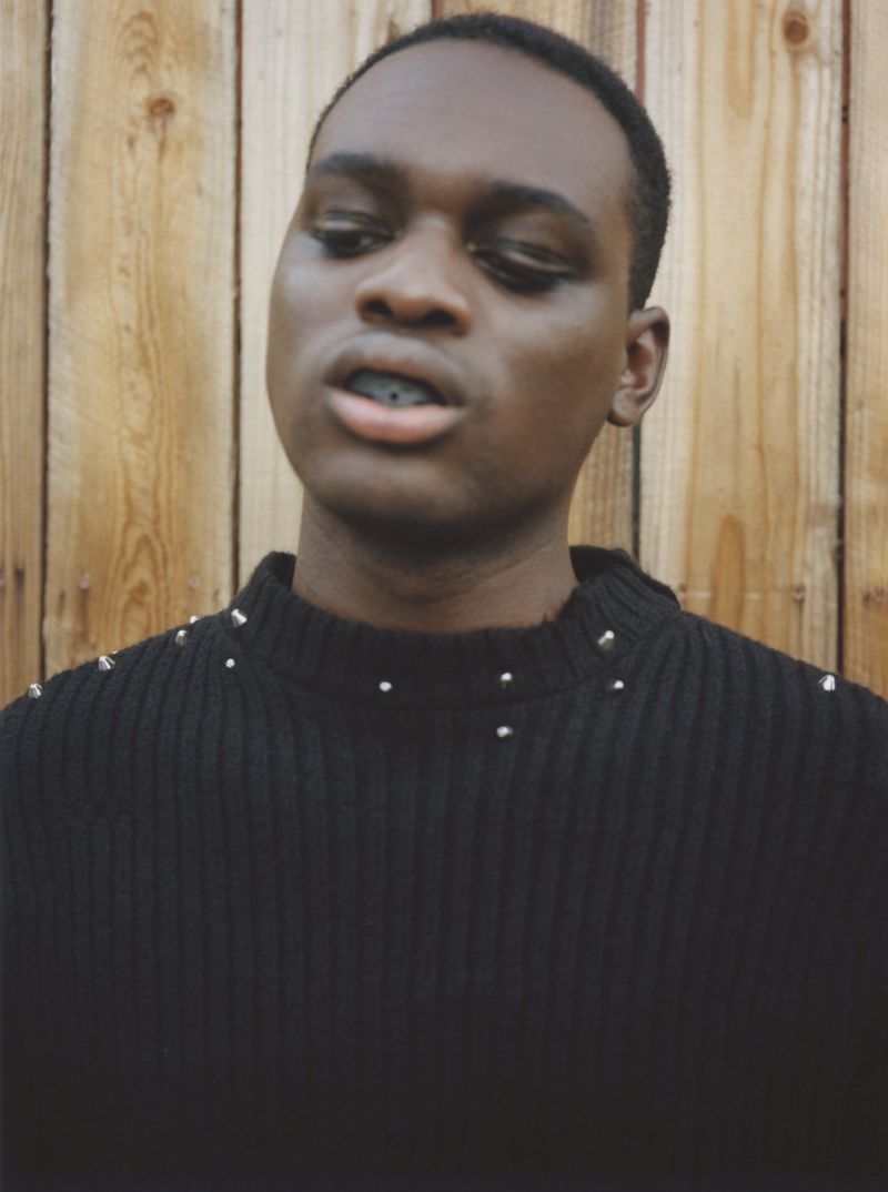 Portrait of a young Black man in a black studded sweater against a rustic wooden background, featuring a close-up of his face and upper body.