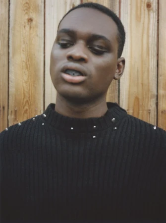 Portrait of a young Black man in a black studded sweater against a rustic wooden background, featuring a close-up of his face and upper body.