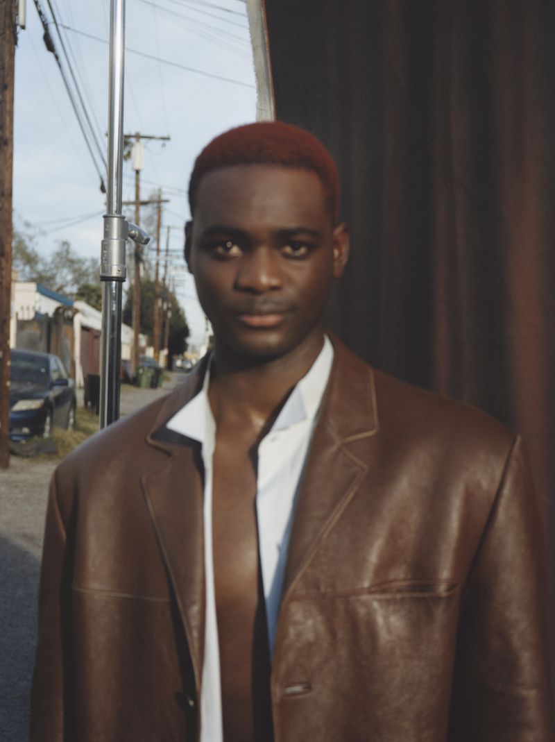 Portrait of a young adult Black man in brown leather jacket and white shirt, street with buildings and telephone poles in background.