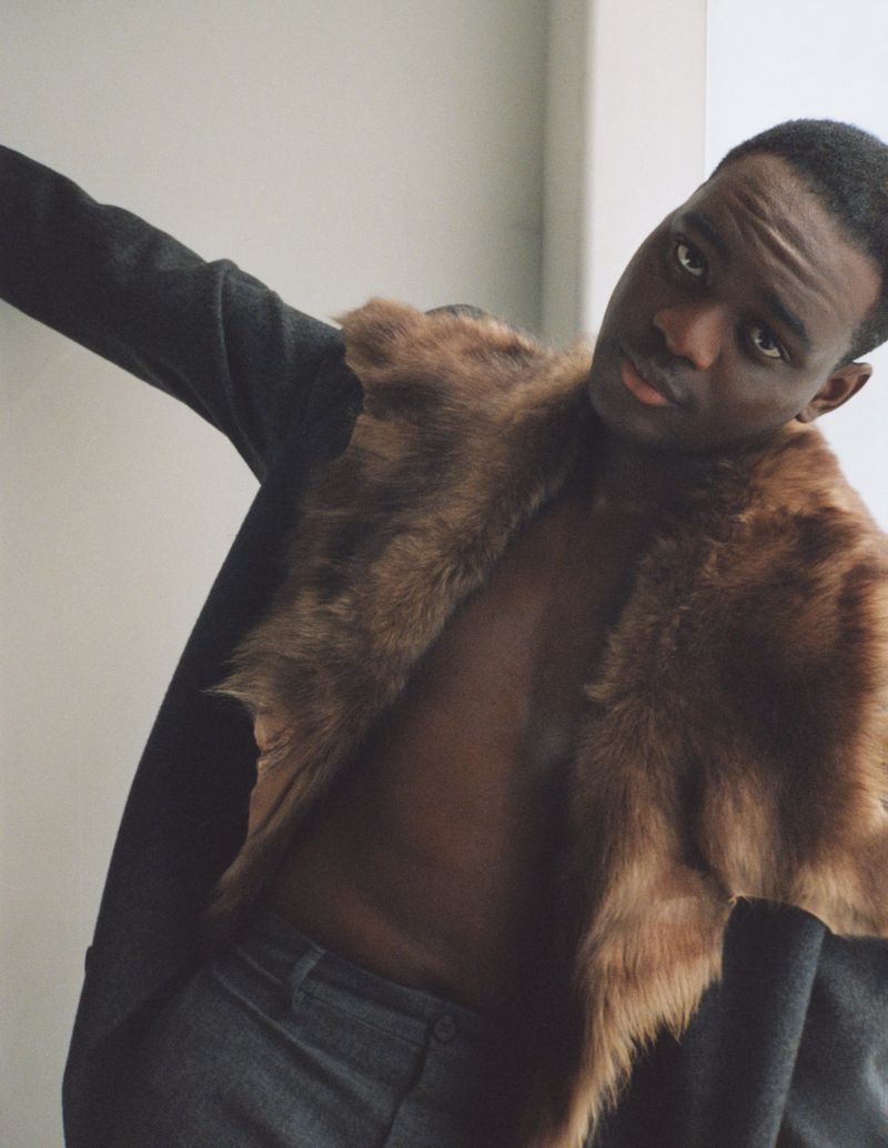 A black man poses in a dark coat with a fur collar and dark pants against a neutral background, creating a striking fashion portrait.