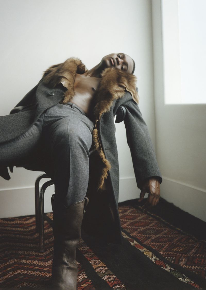 A Black man sits with his eyes closed, wearing grey slacks, a fur-trimmed coat, and brown boots in a neutral setting.