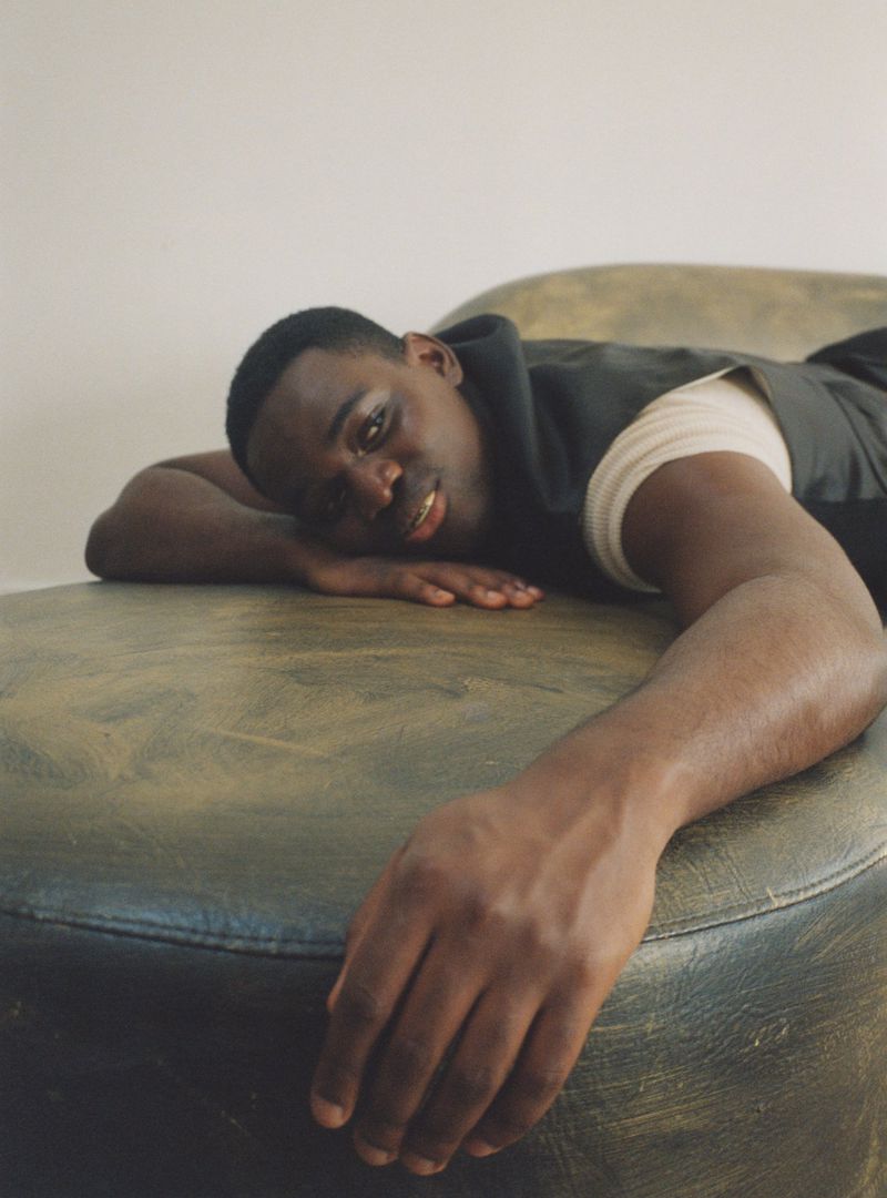 Portrait of a Black man lying horizontally on a green upholstered surface, looking toward the viewer in a relaxed pose, indoors.