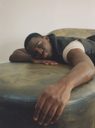 Portrait of a Black man lying horizontally on a green upholstered surface, looking toward the viewer in a relaxed pose, indoors.