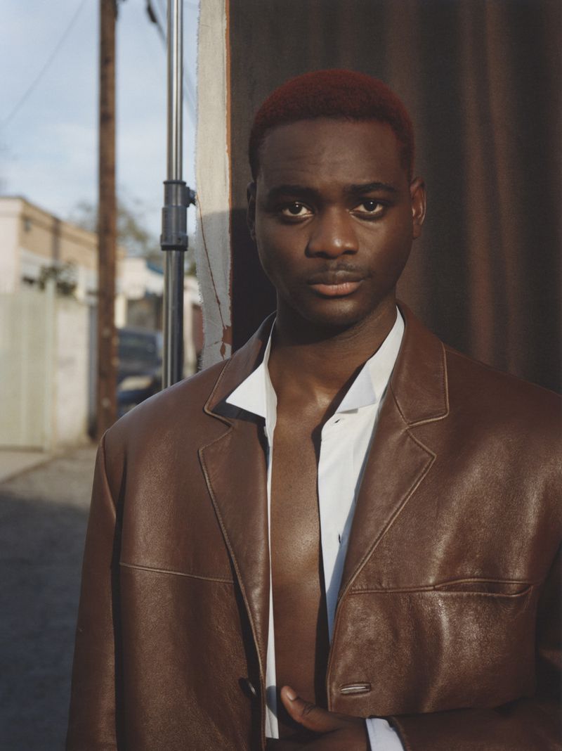 Portrait of a young, dark-skinned male model in a brown leather jacket and white shirt posing in an urban alley setting with soft light.