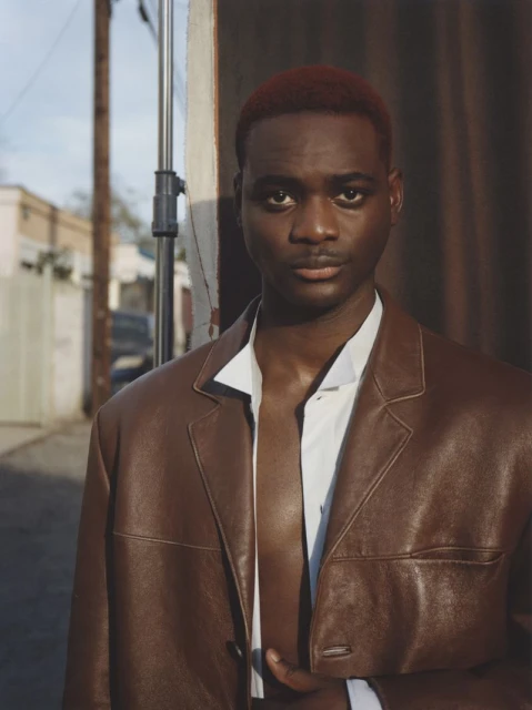 Portrait of a young, dark-skinned male model in a brown leather jacket and white shirt posing in an urban alley setting with soft light.