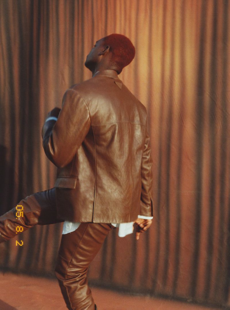 An African male model in a brown leather suit poses indoors in front of a draped brown curtain. He looks up, showing off his red-dyed hair.