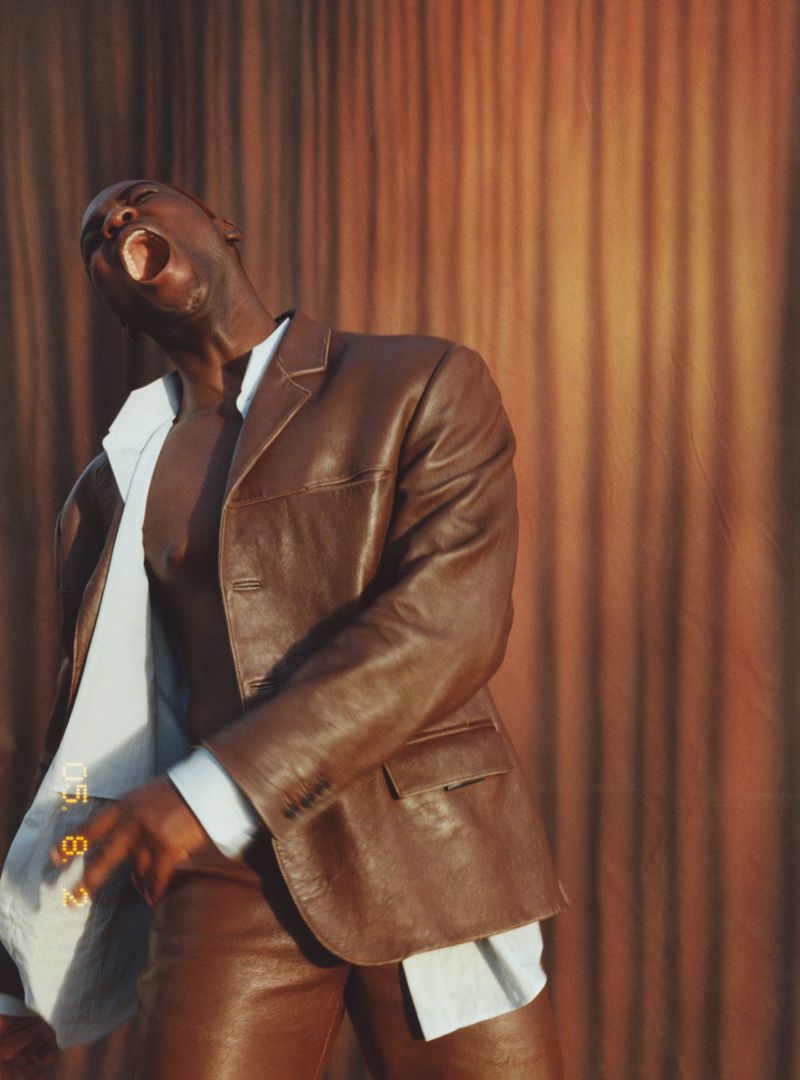A Black male fashion model poses with an open mouth, wearing a brown leather jacket and pants, with a light blue shirt in the background.