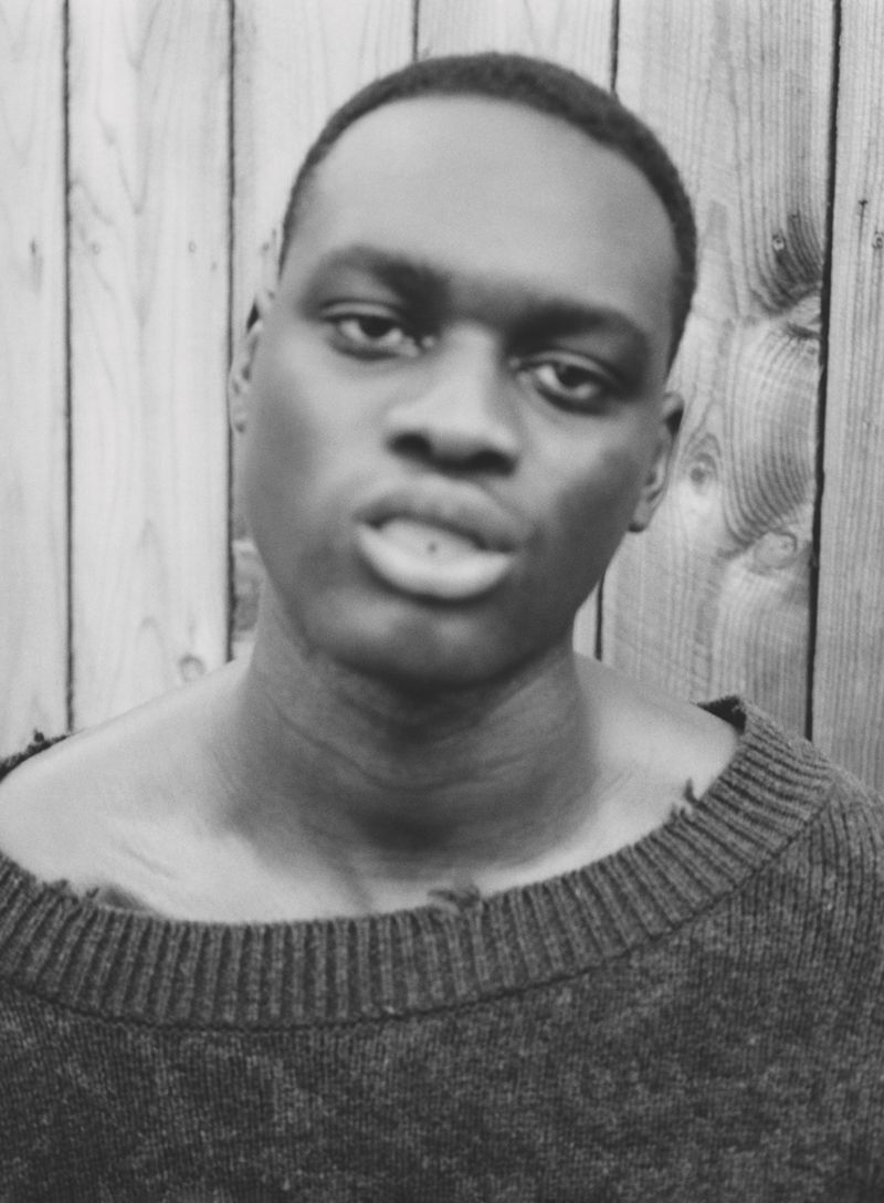 Monochromatic portrait of a Black man wearing a sweater against a light wooden fence background, showcasing his features and attire.