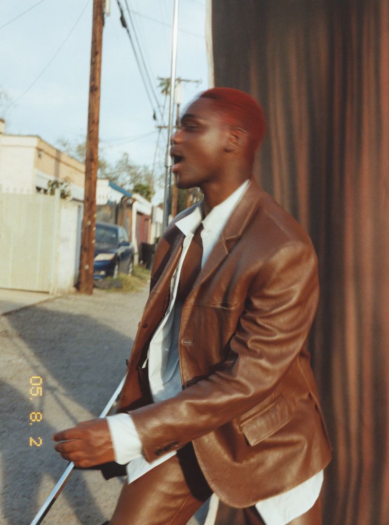 Portrait of African American adult male in brown leather jacket and pants, with red hair, in an outdoor urban setting.