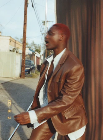 Portrait of African American adult male in brown leather jacket and pants, with red hair, in an outdoor urban setting.