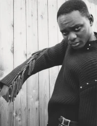 Portrait of a man wearing a dark fringed sweater against a vertical wooden plank fence background
