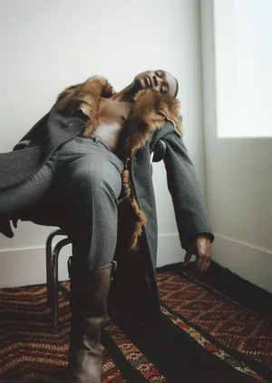 Black man with bare torso wearing a fur-lined coat and boots seated on a chair indoors for AnOther Man Magazine.