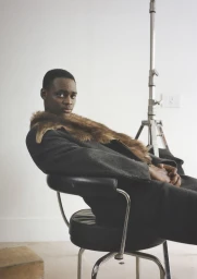 Black man in a dark coat with a fur collar seated in a black chair against a white studio wall with lighting stand
