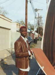 Young Black man in a brown leather suit holding a brown prop in an alleyway for AnOther Man Magazine