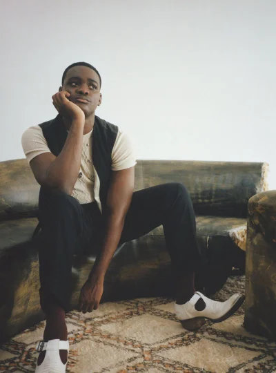 Young Black man seated on a dark curved sofa resting his chin on his hand against a neutral background