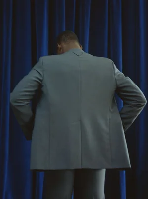 Man wearing a grey suit jacket viewed from behind against blue curtains